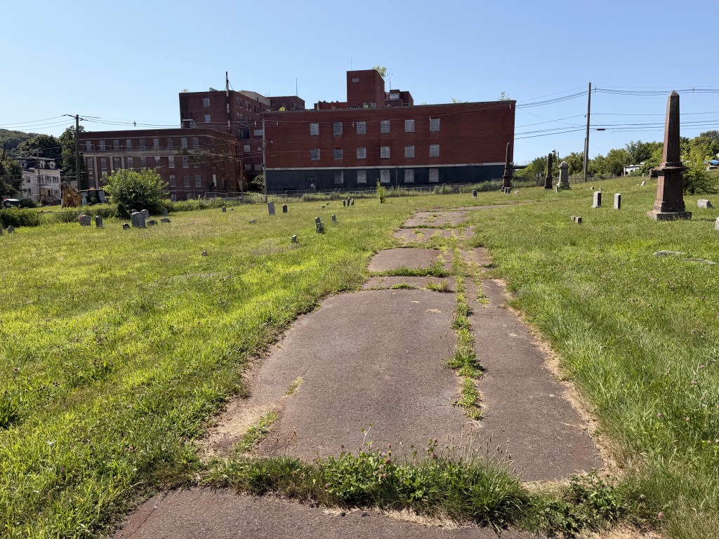 An aging paved path crosses the grass and burial grounds toward a red brick building beyond West Cemetery.